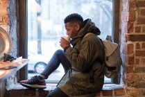 Vista lateral del turista sentado en el alféizar de la ventana en la cafetería y beber bebidas calientes - foto de stock