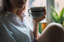 Обрізати жінку в сорочці з чашкою за вікном — стокове фото