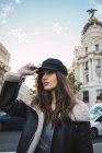 Portrait of brunette woman in cap posing on street and looking away — Stock Photo