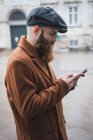 Side view of bearded man in coat and cap using phone on street — Stock Photo