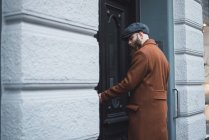 Seitenansicht eines nachdenklichen bärtigen Mannes in Mantel und Mütze, der die Eingangstür öffnet — Stockfoto