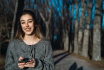 Donna sorridente in maglione che tiene il telefono e ride della macchina fotografica sul vicolo albero autunnale . — Foto stock