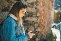 Side view of stylish girl using smartphone on background of stone wall — Stock Photo