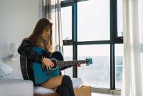 Jovem mulher sentada na cama e tocando guitarra em casa — Fotografia de Stock