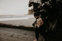Seitenansicht eines hemdslosen Mannes, der sich am Strand auf einen umgestürzten Stamm stützt — Stockfoto