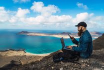 Вид сбоку человека, сидящего на прибрежной скале с ноутбуком на коленях и просматривающего смартфон — стоковое фото