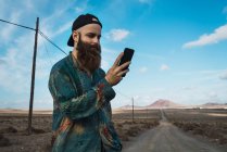 Портрет бородатого чоловіка, який використовує смартфон на дорозі в сільській місцевості — стокове фото