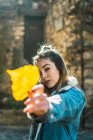 Portrait of woman in jacket outstretching hand with bright yellow leaf looking at camera in sunlight. — Stock Photo
