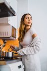Vue latérale de la jeune femme agitant dans le pot et regardant au-dessus de l'épaule sur la cuisine à la maison . — Photo de stock