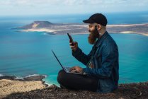 Вид збоку чоловіка, що сидить на береговій скелі з ноутбуком на колінах і переглядає смартфон у сонячний день — стокове фото