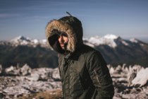 Portrait man in warm coat with hood standing in sunlight on background of snowy mountains. — Stock Photo