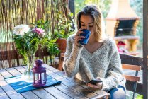 Junge Frau im Pullover trinkt Kaffee, während sie zu Hause Smartphone schaut — Stockfoto