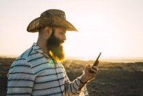 Vista lateral do homem barbudo no smartphone de navegação de chapéu — Fotografia de Stock