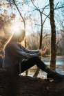 Вид збоку білявка дівчина сидить на стовбурі дерева і приймає селфі в осіннє сонячне світло — стокове фото