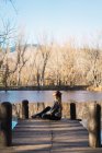 Side view of dreaming woman sitting on wooden pier with lake in fall time on background. — Stock Photo