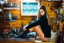 Side view of young woman in sweater and socks sitting on counter in small kitchen and posing sensually. — Stock Photo
