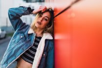 Confident blonde girl in denim leaning on red wall and looking at camera while touching head. — Stock Photo