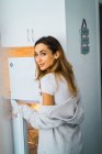 Ritratto di ragazza bruna che apre il frigorifero a casa e guarda dietro le spalle la macchina fotografica — Foto stock