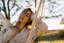 Beautiful young model in sweater posing in hammock on background of countryside. — Stock Photo