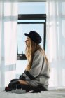 Vista lateral da jovem mulher sonhadora em suéter e chapéu sentado na cama branca com câmera contra janela . — Fotografia de Stock