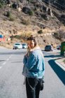 Visão traseira da mulher casual em casaco de ganga na estrada olhando para a câmera sobre o ombro e sorrindo . — Fotografia de Stock