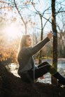 Side view of woman sitting on tree trunk and taking selfie with smartphone in fall woods. — Stock Photo