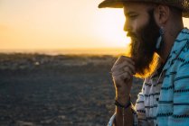 Seitenansicht eines nachdenklichen bärtigen Mannes mit Accessoires und Cowboyhut, der im Sonnenuntergang am Strand posiert. — Stockfoto