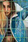 Portrait of provocative blonde girl in denim looking at camera and standing behind metal net. — Stock Photo
