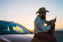 Side view of bearded traveler sitting on car and browsing smartphone — Stock Photo