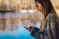 Side view of woman on autumnal pier above pond browsing smartphone — Stock Photo