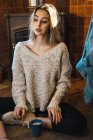 Jovem modelo sensual em suéter tendo xícara de café enquanto posando no chão da casa e olhando para longe . — Fotografia de Stock
