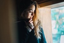 Side view of young woman cuddling in sweater and looking away in window. — Stock Photo