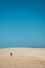 Back view of traveler with backpack walking on endless sandy terrain under bright blue sky. — Stock Photo