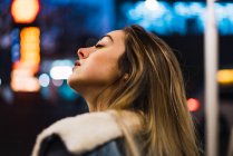 Young blonde girl posing dreamily with eyes closed on background of street night lights. — Stock Photo