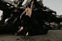 Visão traseira do homem sem camisa pulando na praia com troncos de árvore caídos — Fotografia de Stock