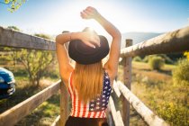 Visão traseira da mulher vestindo chapéu e camisa com EUA bandeira impressão posando na ponte de madeira — Fotografia de Stock