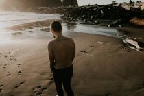 Visão traseira do homem sem camisa em pé na praia ensolarada no oceano . — Fotografia de Stock