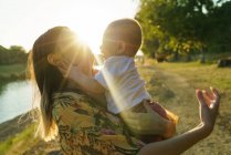 Vista lateral da mãe com a criança a mãos no parque iluminado pelo sol — Fotografia de Stock