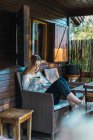 Side view of young woman reading book in armchair at chalet porch — Stock Photo