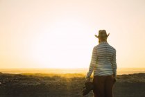 Rear view of man in hat carrying boots in hand and standing at sunset beach — Stock Photo