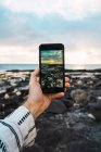 Crop male hand taking shot on smartphone of amazing ocean shoreline with rocky beach. — Stock Photo