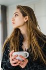 Retrato de menina com cabelo comprido segurando caneca de café em casa e olhando para longe . — Fotografia de Stock