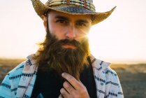 Retrato de homem barbudo em chapéu palmando barba e olhando para a câmera — Fotografia de Stock