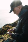 Seitenansicht eines Mannes, der auf einem grünen Hügel steht und am Meer sein Smartphone benutzt. — Stockfoto