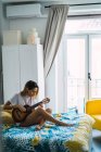 Vista laterale di giovane donna che suona la chitarra a letto a casa — Foto stock