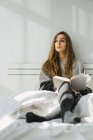 Chica relajante con libro en una cama acogedora y mirando hacia otro lado - foto de stock