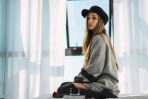Stylisches junges Mädchen mit Mütze sitzt auf dem Bett und schaut weg — Stockfoto