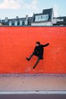 Vista lateral del hombre en ropa de abrigo saltando en la pared naranja en la calle . - foto de stock