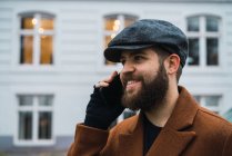 Sonriendo hombre barbudo hablando en el teléfono inteligente en la calle - foto de stock