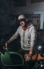 Portrait of man in cap posing with custom motorbike at workshop — Stock Photo